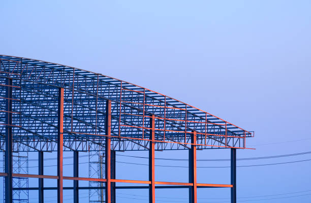 Orange sunlight reflection on surface of metal industrial warehouse building with dome roof structure in construction site against blue evening sky background at sunset time